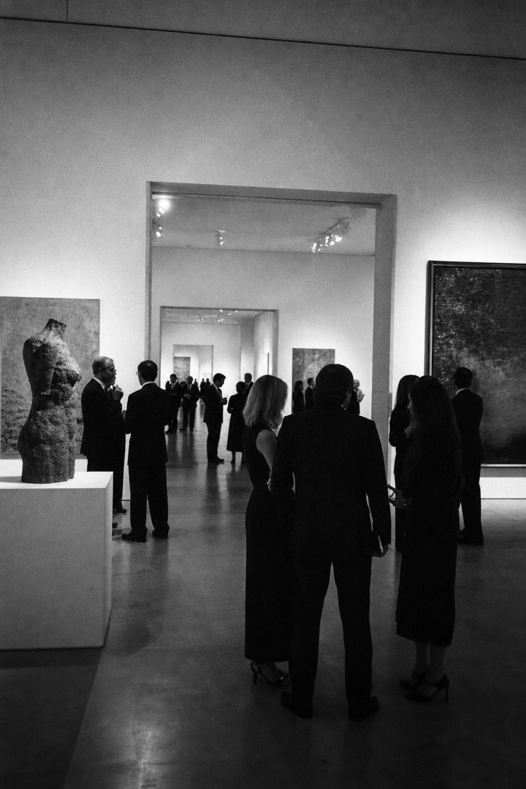 Art dealers and gallerists in quiet conversation at a private evening in a contemporary gallery, rooms opening onto further rooms
